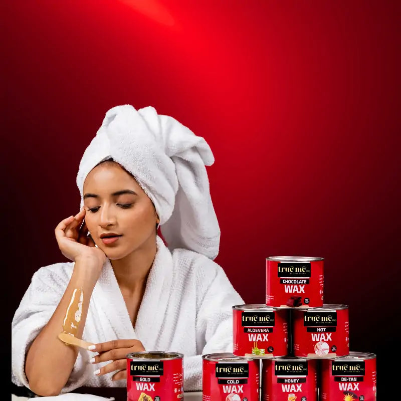 Eva Giri with a towel on their head sitting in front of a stack of True Me Hair Wax containers, with wax applied to their leg.